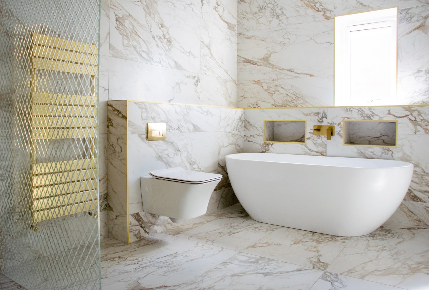 A marble-look bathroom with a freestanding bath, a wall-hung toilet and a brass heated towel rail