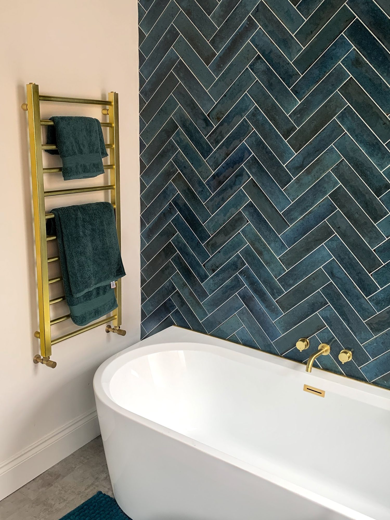A freestanding bath beside a wall of teal herringbone tiles and a brass heated towel rail